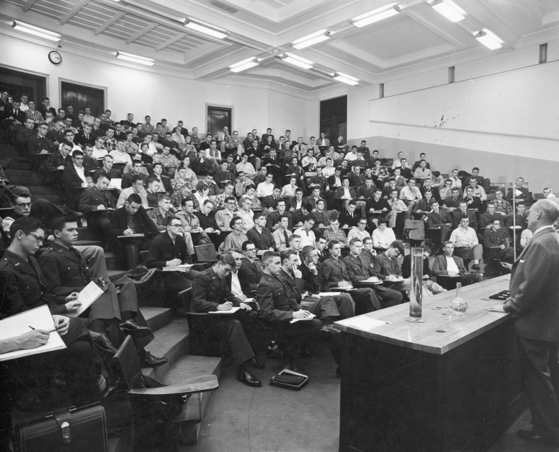 A chemistry lecture class being conducted in the stadium lecture hall within the Chemistry Building (now Gilman Hall). A lecturer stands behind a long demonstration counter at the front of the class. This photographic view is from the left side of the lecturer looking toward the lecture audience.