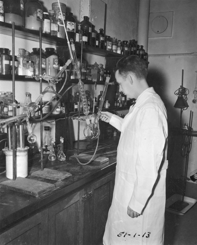 A chemistry student at work in a laboratory. Dressed in a white lab coat, the student is standing before a distillation apparatus with a wall of reagents on shelves behind the equipment.