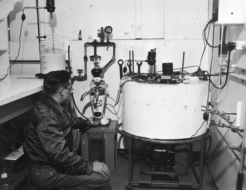 G. F. Hanna, a graduate student in the Department of Chemistry and a research fellow of the Agricultural Experiment Station, showing the refrigerated super centrifuge along with the refrigerated bath. The room was maintained at a temperature just above freezing and was used in preparation and fractionation of proteins, enzymes and other materials of biochemical importance that are frequently unstable at room temperature. The refrigerated bath provided even lower temperatures that might have been required for certain steps in processing. The super centrifuge rotated at speeds up to 50,000 rpm permitting separations not possible in more conventional centrifuges. Materials undergoing centrifugation could be maintained at low temperatures by circulation of coolant through coils from the refrigerated bath. Research conducted in this facility was sponsored primarily by the Agricultural Experiment Station. It was located in the Biophysical Chemistry Laboratories in Chemistry Hall (now Gilman Hall). This image is very similar to no. 1052-3-3.