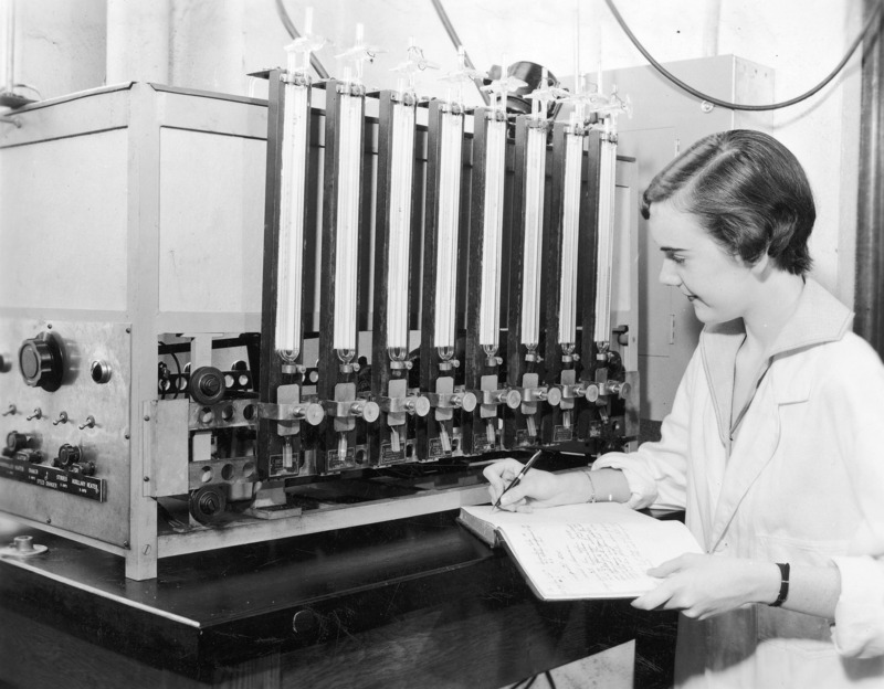 A female student in a white lab coat using a Warburg apparatus. The Warburg apparatus was widely used in biochemical and biological research for measuring rates of reaction in which gases are involved. The student is noting readings in a lab record book.