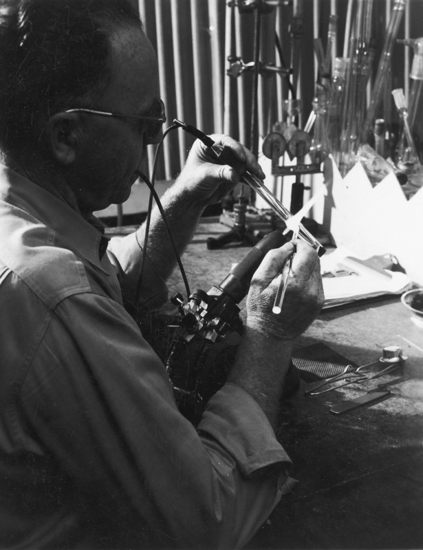 A technician hand-making a piece of laboratory glassware with a torch for heat. Holding a piece of glass rod stock in the torch with one hand, the technician is modifying the glass tube by touching it with a glass rod in the flame. Glass lab ware hangs on the wall behind the technician's work bench.