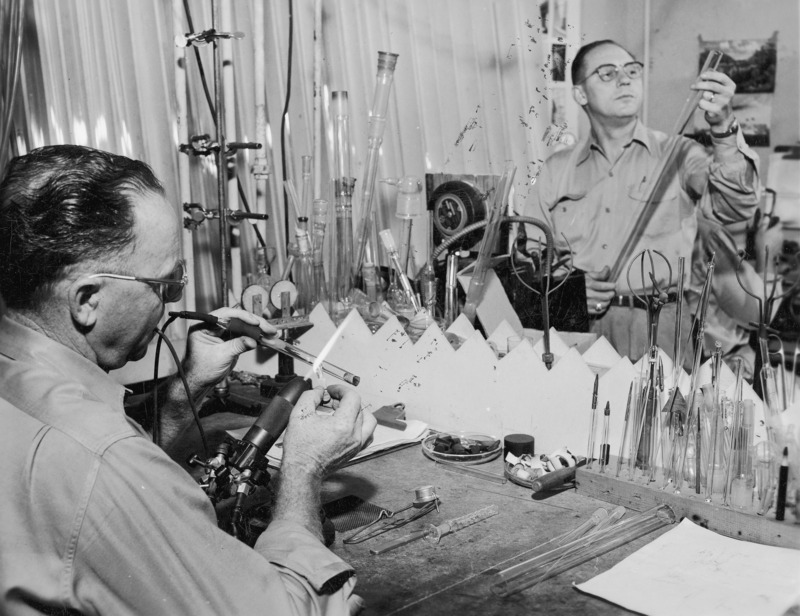 Two technicians in the glass lab of the Chemistry Department. One technician is working with a torch and tubes of glass at a worktable, on which are a lighter, glass tubes, rubber stoppers and various tools.The other technician is standing in the background inspecting a large glass tube.