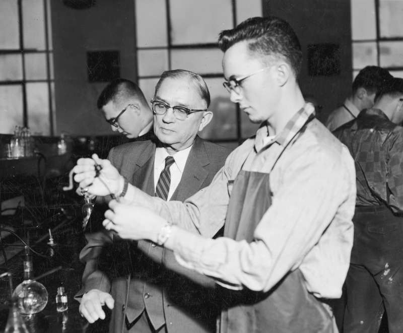 Professor F. E. Brown observing a chemistry student in the lab. The student is using a dropper to add liquid to a test tube. Three other students are working in the background.