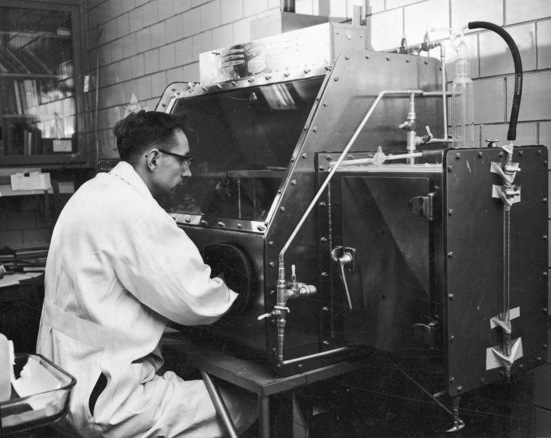 Jim Weiher at the dry box (a shielded laboratory chemical containment hood), which was used to perform chemical operations in a controlled atmosphere. Weiher is seated, with his arms inserted through portals into the inner chamber which can be viewed through a glass cover.