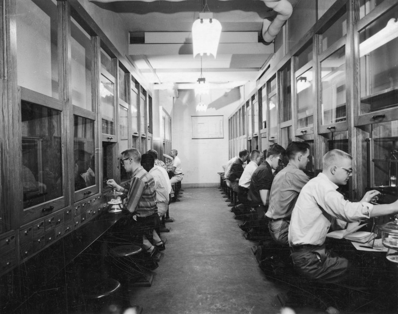 The balance room used for quantitative analysis. Contiguous fumehoods line both walls of a long narrow room, down the center aisle of which are arranged chairs at each fume hood work station. Sitting back to back in two rows, students are occupied in their individual experiments.