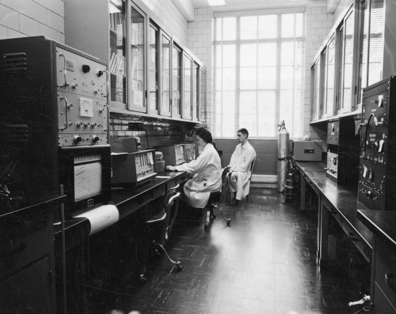 The counter room for measuring radioactivity. Two technicians, a man and a woman,are seated at the far end of the room; the woman is working with one of the machines. A number of pieces of equipment are shown.