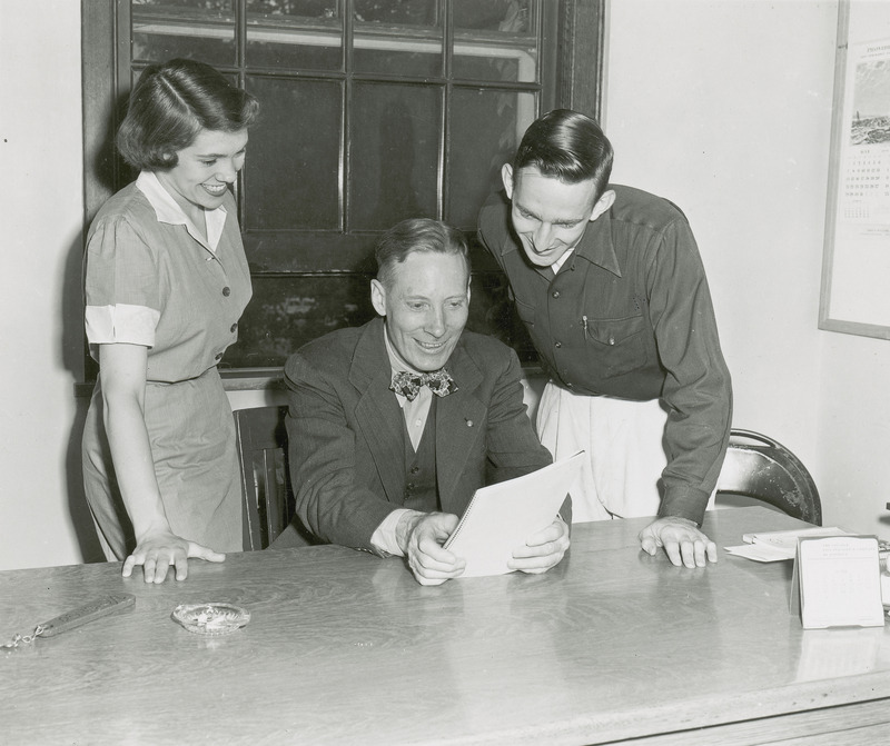 Red Barron is seated at a desk and is flanked on either side by two standing individuals. These two individuals are members of the Homecoming Central Committee. All three are reading a document which Barron is holding.