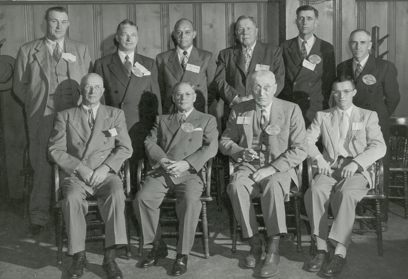 Ten alumni are arranged in two rows for this formal portrait. The front row is seated while six individuals stand behind them. Most are wearing paper name tags and a few have football shaped tags indicating membership on the football team.