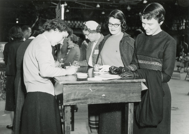 Four women are standing at a table while filling out Homecoming registration cards. Other individuals are in the background.