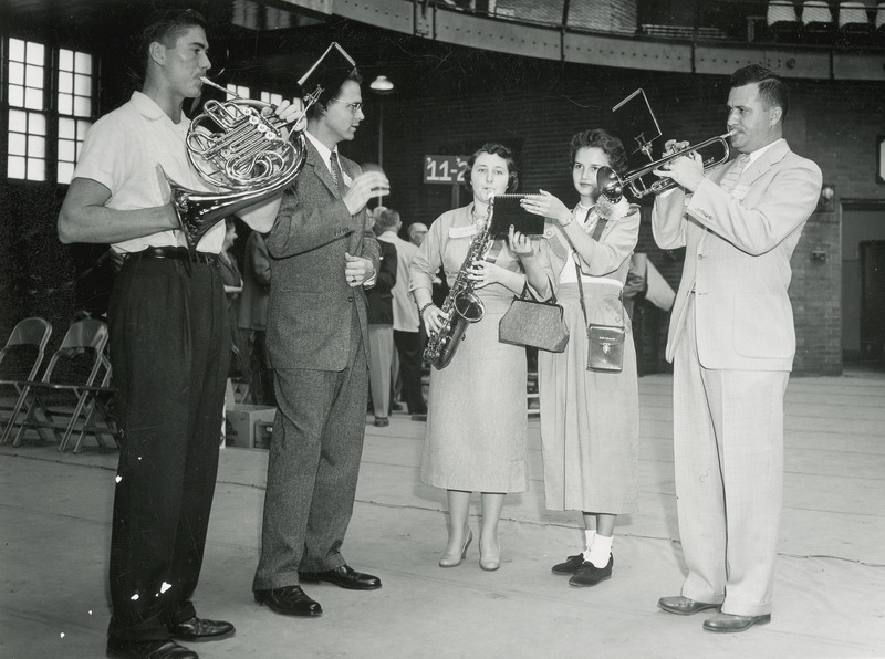 Five members of the Alumni band are performing at a Homecoming event. Three individuals are playing instruments (trumpet, saxophone and French horn), one is conducting and the other is holding music.