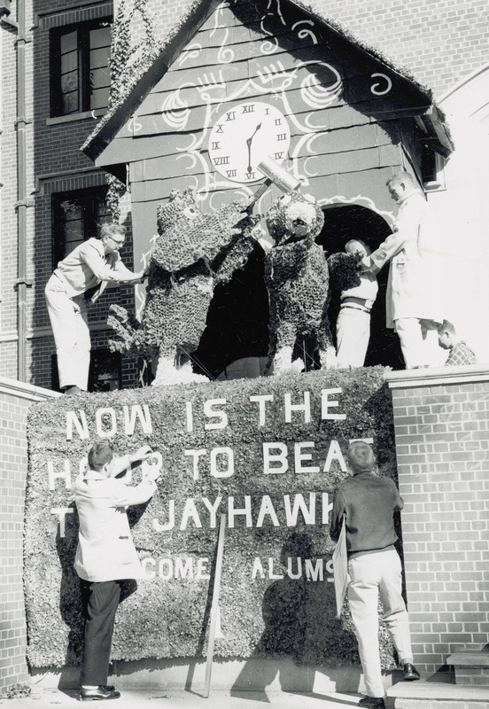 The Birch Hall, Stevenson House Homecoming display of "Now is the Hour to Beat the Jayhawks" won first place in the Men's Residence Association, 1958. Students are shown working on the display, located in front of Friley Hall. It features a large cuckoo clock in which Cy strikes the Kansas University Jayhawk when both emerge from the clock.