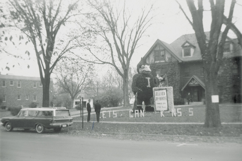 A large ISU mascot, Cy is standing next to a can labeled, "Jellied Jay-birds Canned by ISU, Inc." The upper portion of a Jayhawk, the Kansas University mascot, is in the can. A sign in front of the display reads, "Lets can Kansas.".