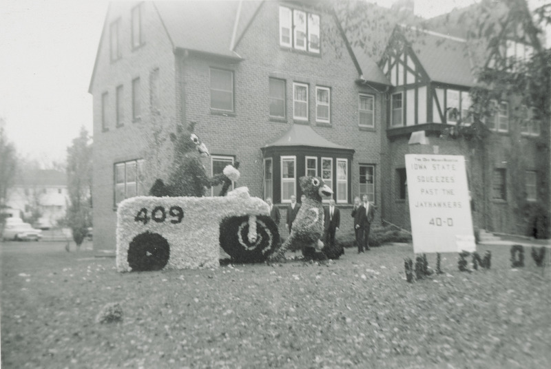 Cy, the ISU mascot is driving a steam roller over the tail of the Kansas University mascot, Jayhawk in this lawn display from Theta Xi, Mu Chapter. A sign nearby partially reads, "Iowa squeezes past Jayhawks 40-0." A group of men in suits are in the background looking at the display.