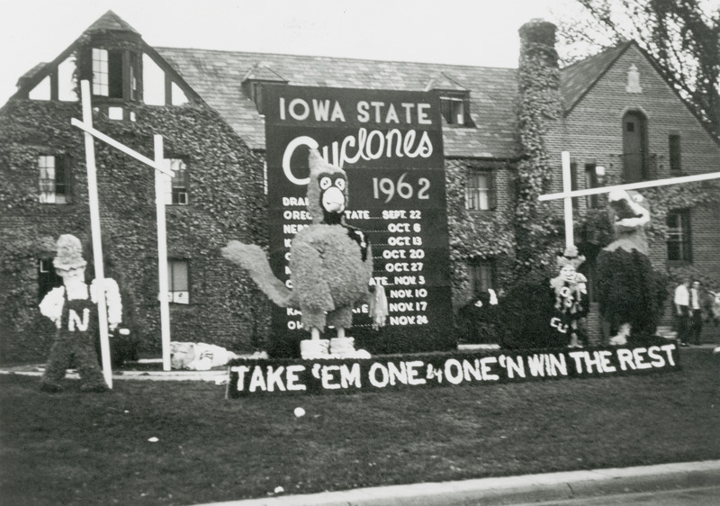 Cy is standing between two football goal posts and is surrounded by smaller figures representing the mascots of other football teams. In back of Cy is a large poster of the football game schedule for 1962. In front of Cy is a sign, "Take 'em one by one 'n win the rest.".