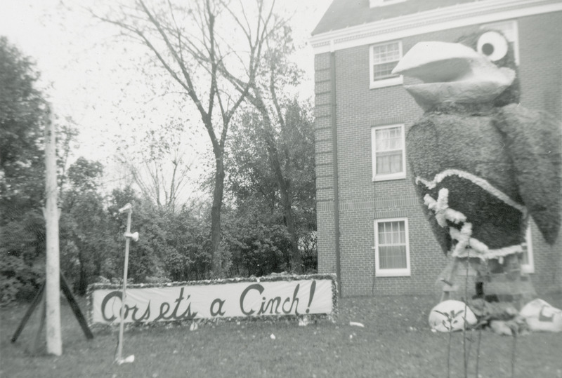 A Kansas University Jayhawk figure is dressed in a tightly laced corset and shirt made of sheer fabric. The sign beside the Alpha Gamma Delta, Rho Chapter's display reads, "Corset's a cinch.".