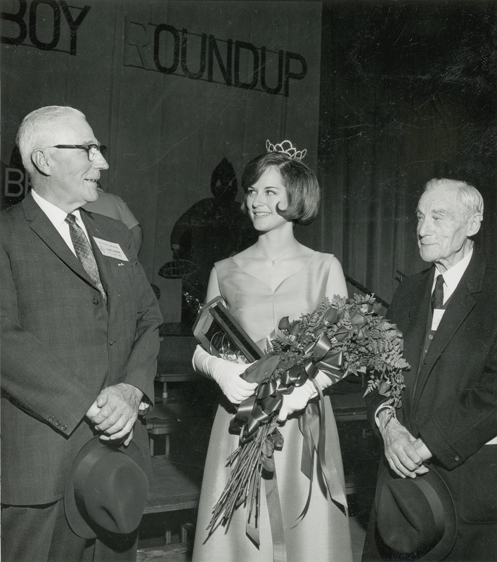 Homecoming queen Adele Berry is standing between to older gentlemen. She is wearing a long formal dress, crown, long with gloves. She is holding a trophy and a bouquet of roses.