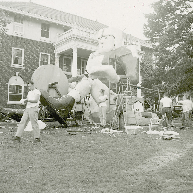 A group of individuals are working on the Beta Theta Pi, Tau Sigma Chapter, lawn display. The display shows a large Iowa State University football player sitting on a depiction of the Kansas State University mascot, the Wildcat. The football player is sharpening a sword on a foot powered grinding wheel.