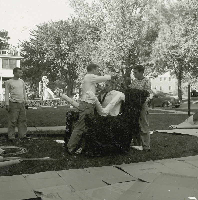 Three individuals are working on the Theta Xi, Mu Chapter, homecoming lawn display of a man seated in an easy chair. On the far side of a short hedge, another group of individuals are constructing a different display.