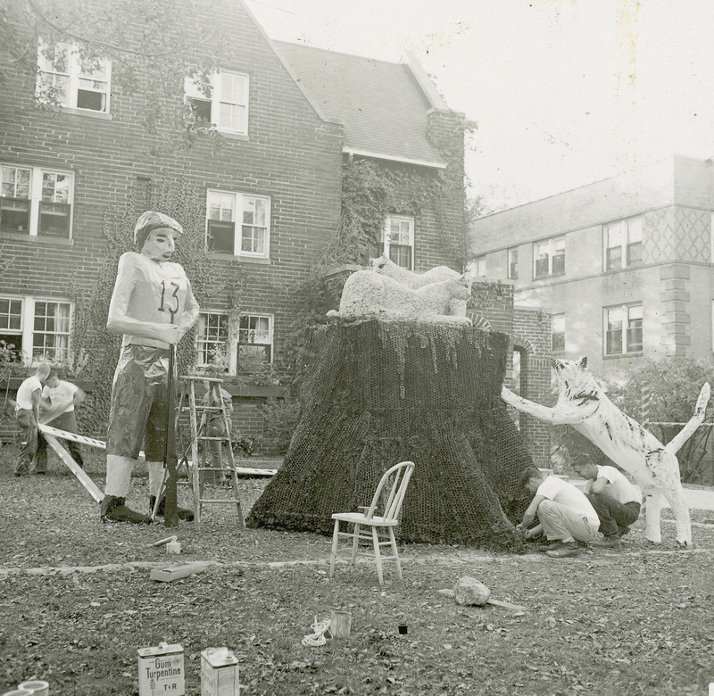 Several members of the Farm House fraternity are working on the House's homecoming lawn display. Included in the display are a standing Iowa State University football player and three cats representing the Kansas State University wildcats. Two cats on top of the stump and a third is propped against the stump side. The football player is carrying a long rife-type object.