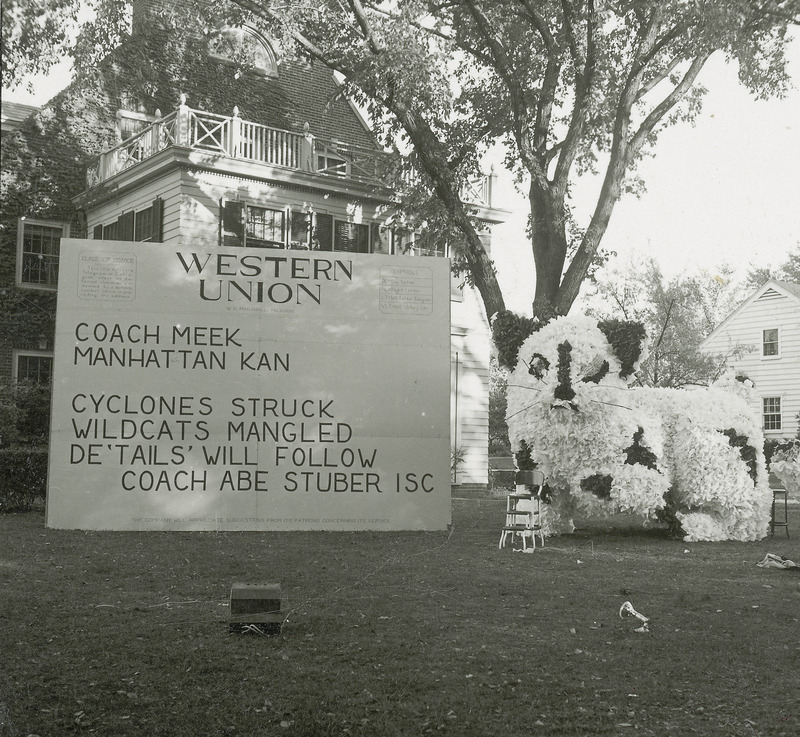 A large cat figure is the central focus of the Delta Delta Delta, Omega Delta Chapter's lawn display. Next to the cat is a sign which is formatted like a telegram reading, "Western Union, Manhattan, Kan., Cyclones struck, wildcats mangled, Details' will follow, Coach Abe Stuben, ISC.".