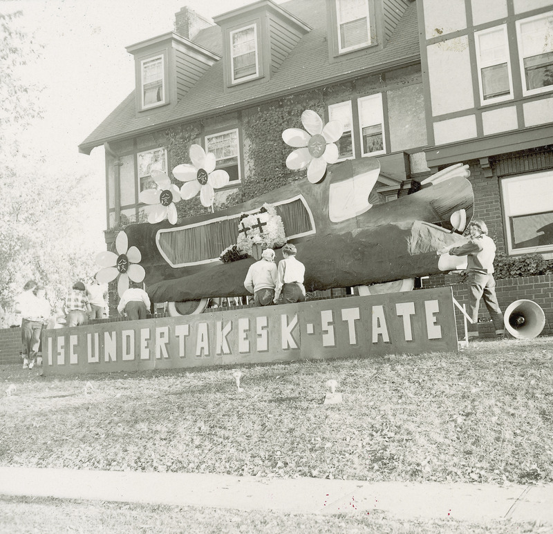 A comic hearse containing a wildcat figure is being constructed by several workers. There are flowers on the hearse roof and a sign in front saying," ISC undertakes K-State.".