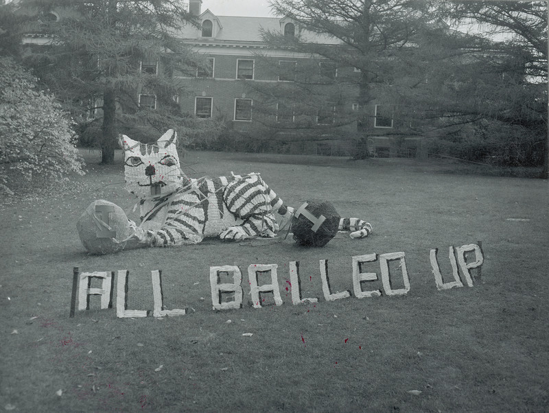 A large cat figure is tangled up in two balls of yarn. The balls of yarn both are labeled with the letter "I." In front of the Lyon residence hall lawn display are the words, " All balled up." each letter is a separate unit.