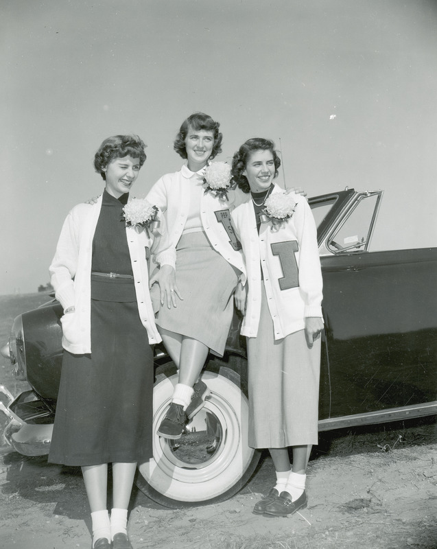 The Homecoming queen and attendants are posed next to a convertible. All of the women are wearing cardigan letter sweaters and mum corsages. They are from left to right: Bonnie Helfrich, Doris Blair (seated, queen) and Ann Bradley.