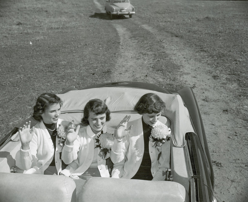 The Homecoming queen and attendants are riding in the back seat of an open convertible. All three women are waving. They are from left to right Bonnie Helfrich, Doris Blair (queen) and Ann Bradley.