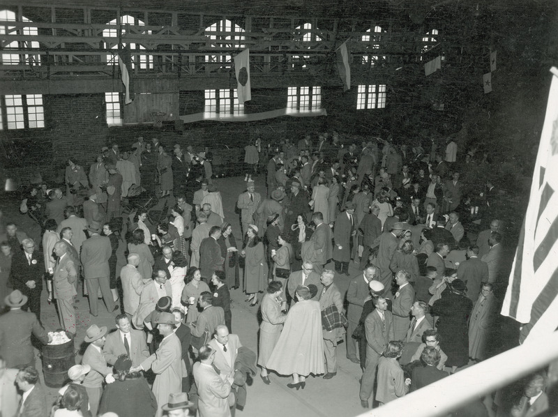 A large group of spectators are gathered in State gym after the football game. Various small groups of individuals are talking or engaged in other activities.