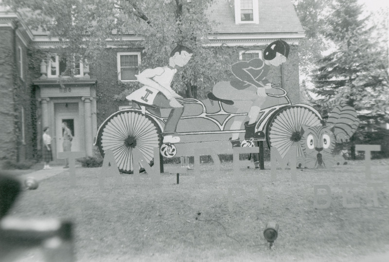 Two cut out figures pedal a tandem bicycle in this Pi Beta Phi lawn display. The front figure is dressed as a football player, while the back figure might be an ISU fan. A tiger figure is being hit by the front of the bicycle.