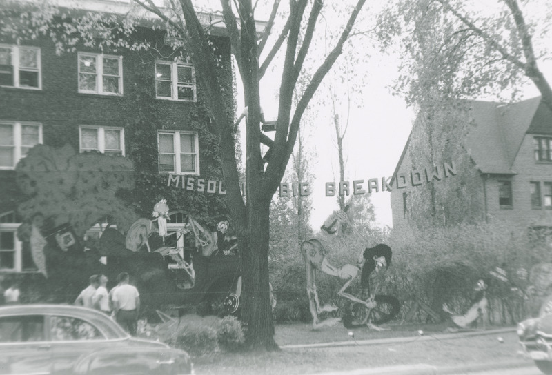 Several individuals are putting finishing touches on this lawn display. The display depicts four bearded cut-out figures dressed in ragged pants who are working to fix a broken-down car. A sign, which is partially obscured by a tree hangs over the display.