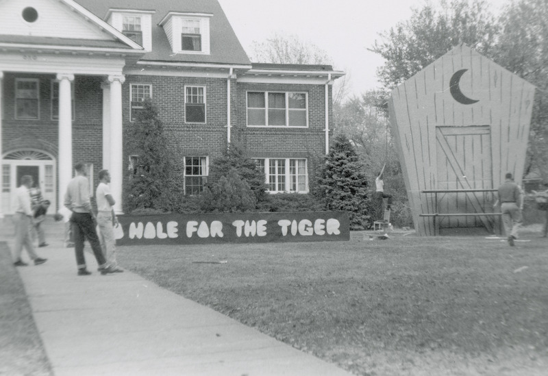 Several individuals are working on this Homecoming lawn display of a large old fashioned out house. Beside the display is a sign that is partially obscured by onlookers. The visible part of the sign reads, ".Hole for the Tigers.".