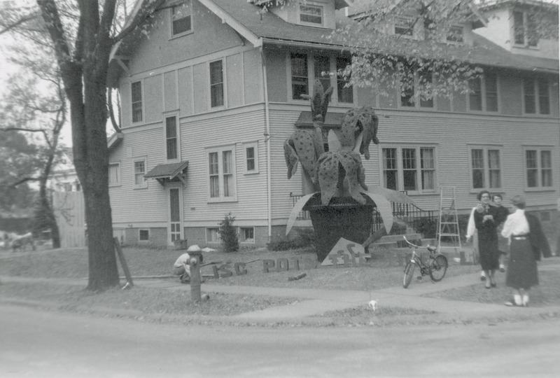 A large potted plant figure is located near the Kappa Alpha Theta, sorority house. A sign in front of the display partially reads, "ISC pots the " A bicycle parked on the sidewalk hides the rest of the sign. A group of women standing near the display are talking to each other.