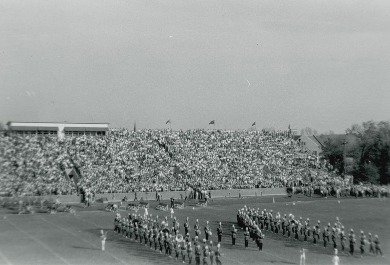 The Iowa State University marching band is performing on the field at the Homecoming football game by forming a giant letter "M. In the background the bleacher seats are full of spectators.