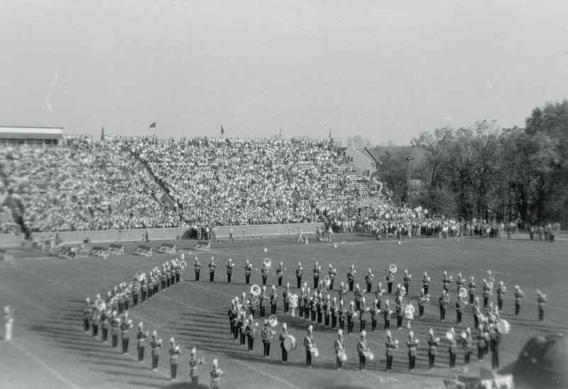 The band is performing marching formations on the field during the Homecoming football game. Behind the band there are spectators seated in the bleachers and standing on the sidelines.