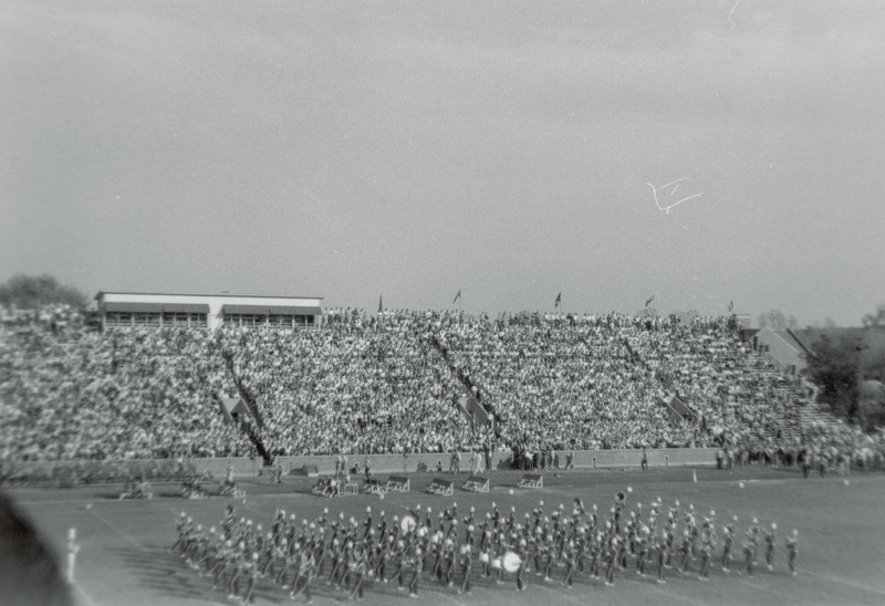 The band is marching lengthwise on the football field in a formation of twelve straight lines. A drum major is facing the first line of band members. A crowd of spectators are in the bleachers.