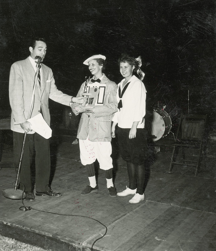 The two member winning team representing Chi Omega, Eta Beta Chapter Sorority appear on stage with WOI-TV personality Joe Adams. Adams, who is the master of ceremonies, is presenting the winners with a trophy. The women are dressed in costumes that resemble late 19th century athletic wear. One women is dresses in bloomers and a sailor middy blouse. The other woman is wearing knickerbockers, a striped sport coat and bowtie.