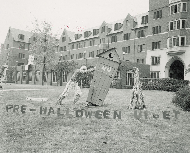 A football player is tipping over an old style outhouse with "MU' written on the door. A long tail type object is coming out from a crack in the from door. It may represent the tail of the University of Missouri's mascot, the Tiger.