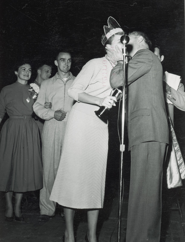 Pat Nelson appears on a stage shortly after receiving a trophy cup. She is being kissed on the check by an unidentified male. Other individuals stand in the background.