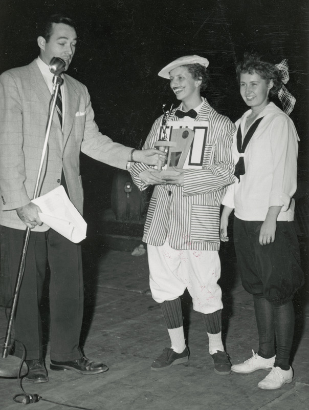 The two member winning team representing Chi Omega, Eta Beta Chapter, Sorority appear on stage with WOI-TV personality Joe Adams. Adams, who is the master of ceremonies, is presenting the winners with a trophy. The women are dressed in costumes that resemble late 19th century athletic wear.