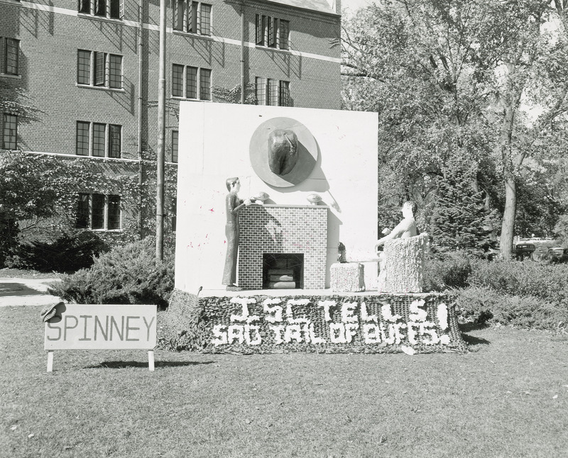 The Spinney House lawn display is modeled on room containing an easy chair and brick fire place. Above the fireplace is rear and tail of a buffalo is mounted like a hunting trophy. On the fireplace mantle are two footballs labeled 54 and 26. One human figure is sitting in the easy chair, while the other is standing.