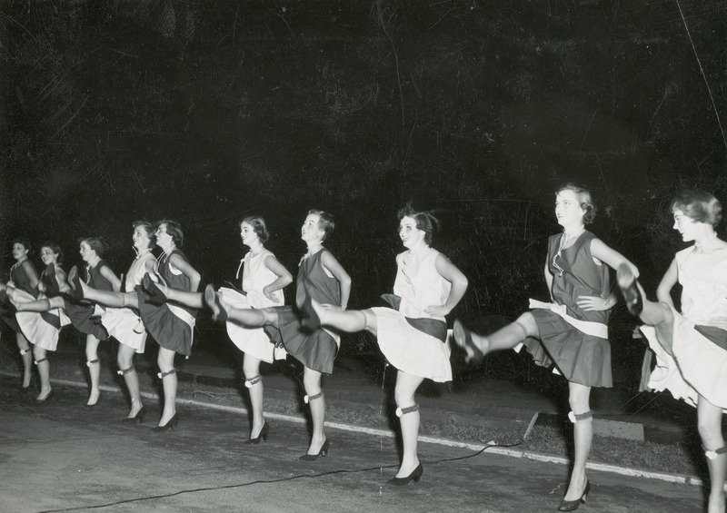 This chorus line consists of ten young women wearing mid-thigh length dresses, high heels, hose and a garter on the left leg below the knee. The women's dresses alternate between light and dark colors. The women are performing during the barbeque entertainment.