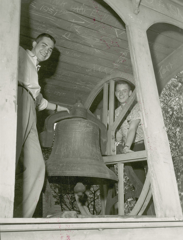 Jerry Whittlesey (left) and Dave Lebuhn (right) pose on opposite sides of the Victory Bell. Many examples of graffiti are visible on the bell and the wooden roof structure that covers it.