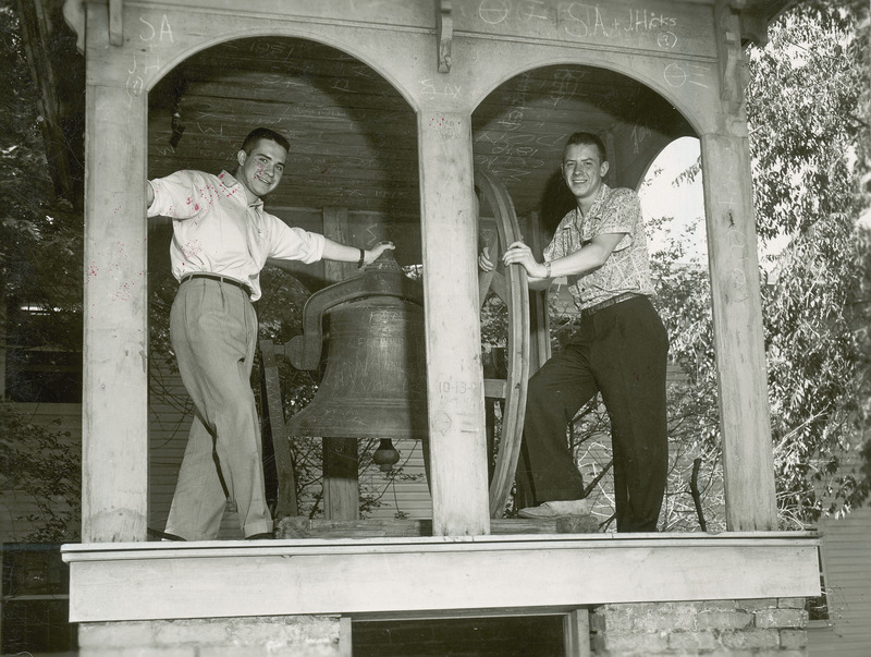 Jerry Whittlesey (left) and Dave Lebuhn (right) pose on opposite sides of the Victory Bell. The two men are framed in the arches of the wooden roof structure that covers the bell.