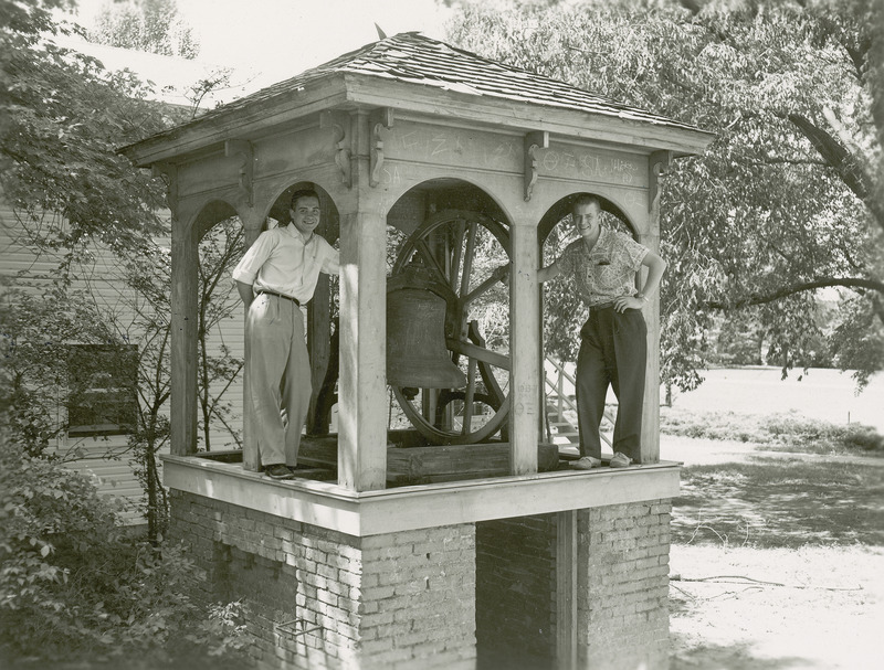 Jerry Whittlesey (left) and Dave Lebuhn (right) pose on opposite sides of the Victory Bell. The brick foundation on which the bell rests and the arched roof structure covering it are visible.