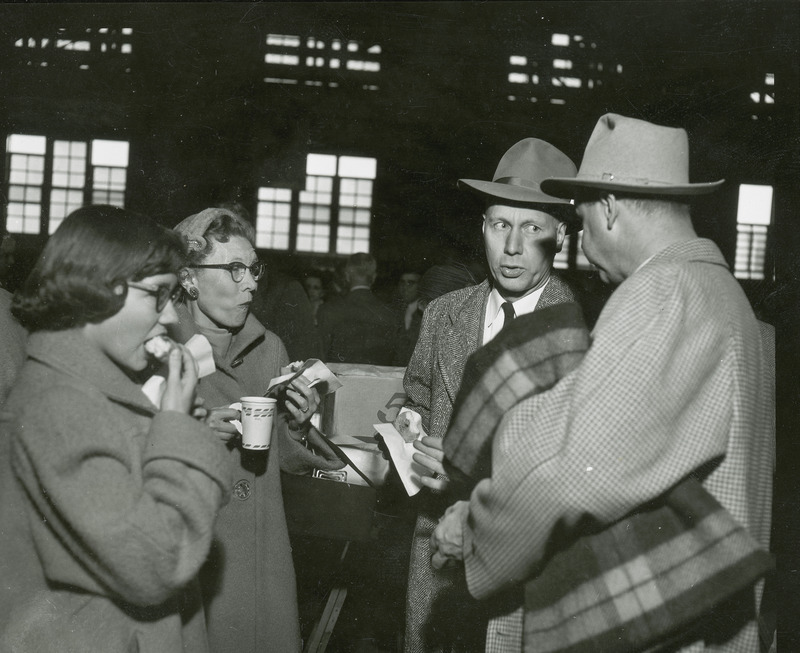 A group of four individuals are eating food and drink beverages at a Homecoming event. All four are wearing coats and one man is carrying a blanket.