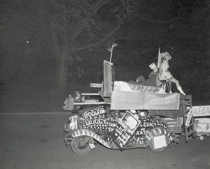 The Phi Kappa Scrap Heap Scramble entry is being driven past a small group of spectators. The car is decorated with paint and has a bed attached to the roof.