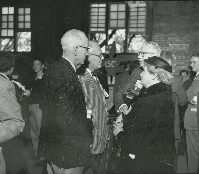 A group of individuals including D. R. "Spec" and Mrs. Collins are enjoying conversation and refreshments at a Homecoming event.