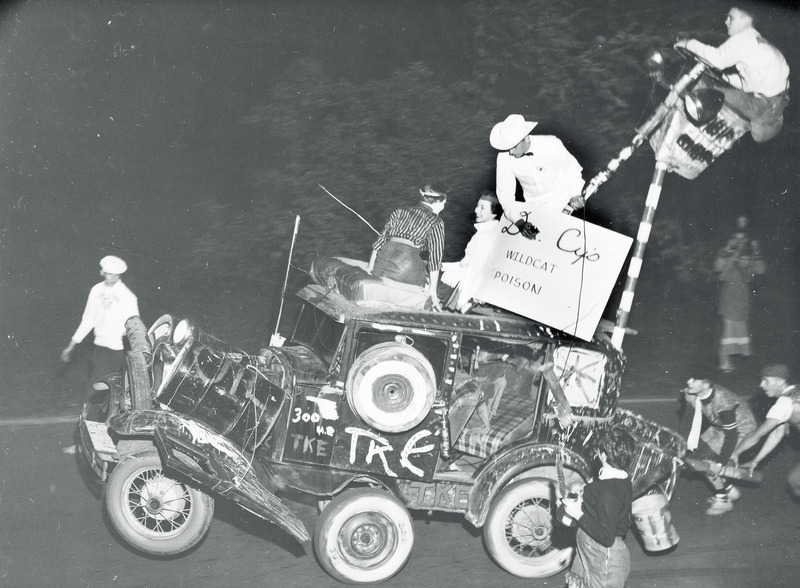 Tau Kappa Epsilon, Epsilon Chapter, entry in the Scrap Heap Scramble is a Ford car with an extra set of wheels mounted in front. Several people are riding on the car, while others push it along the street.