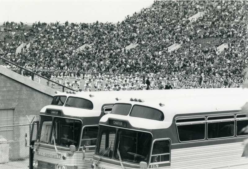 Crowds of spectators are leaving the bleachers at the end of the homecoming football game. Both football teams are also leaving the field. In the foreground, two charter buses wait for passengers.